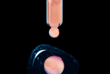 Un compte-gouttes en verre tenant une goutte de liquide rose au-dessus d'une surface bleue brillante.