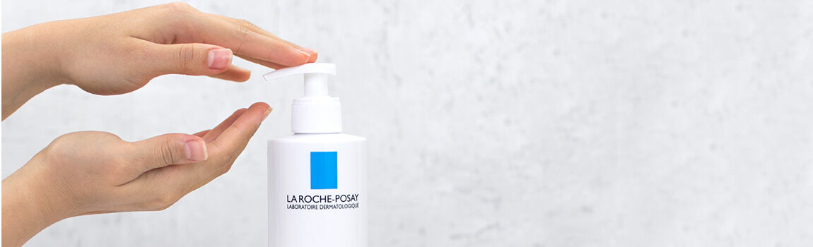 A person’s hands are positioned above a white pump bottle with a blue label, ready to dispense a product from it. The background is a light, textured surface.