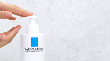 A person’s hands are positioned above a white pump bottle with a blue label, ready to dispense a product from it. The background is a light, textured surface.