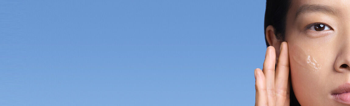 A close-up of a person's face against a blue background, applying a skincare product to their cheek.