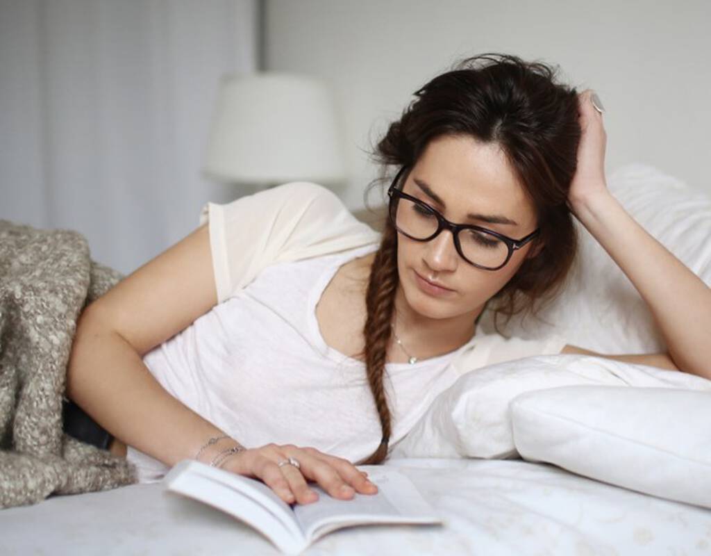 Une jeune femme en train de lire un livre, allongee sur un lit, avec des lunettes. Elle a les cheveux en tresse et porte un t-shirt decontracte.