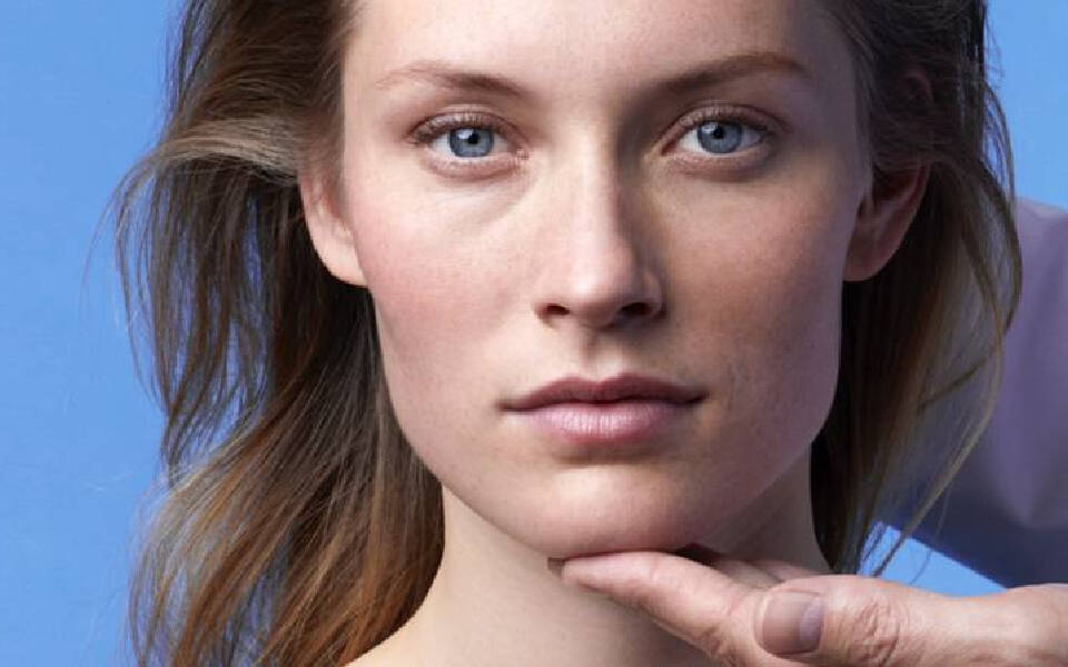 Portrait d'une femme aux cheveux laches, avec une main posee delicatement sous son menton, sur fond bleu clair.