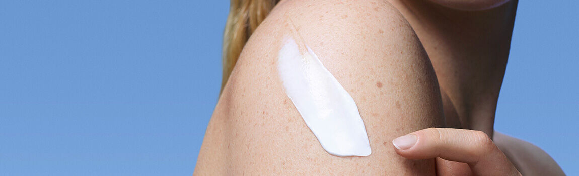 A close-up of a woman's shoulder with a creamy lotion being applied, set against a clear blue background.