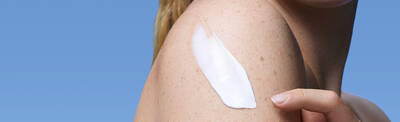 A close-up of a woman's shoulder with a creamy lotion being applied, set against a clear blue background.