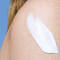 A close-up of a woman's shoulder with a creamy lotion being applied, set against a clear blue background.
