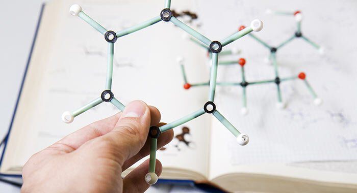 A person's hand holds a molecular model made of colored plastic sticks and balls, with a chemistry textbook open in the background.