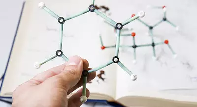 A person's hand holds a molecular model made of colored plastic sticks and balls, with a chemistry textbook open in the background.