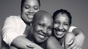 Three women of diverse appearances embrace each other, smiling joyfully against a gray background.