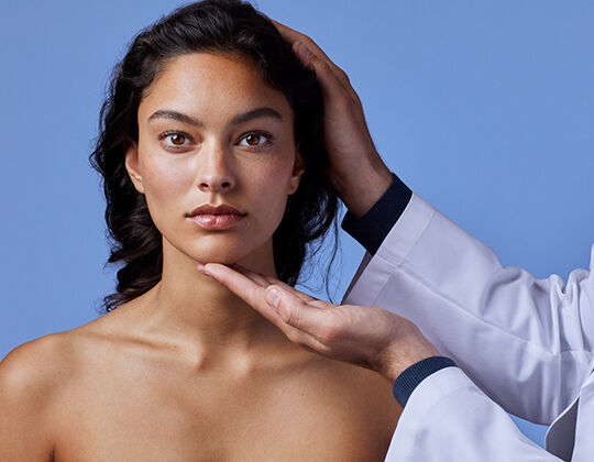 A close-up of a woman's face with long dark hair, showcasing her natural beauty, as a hand gently touches her chin. The background is a soft blue color.