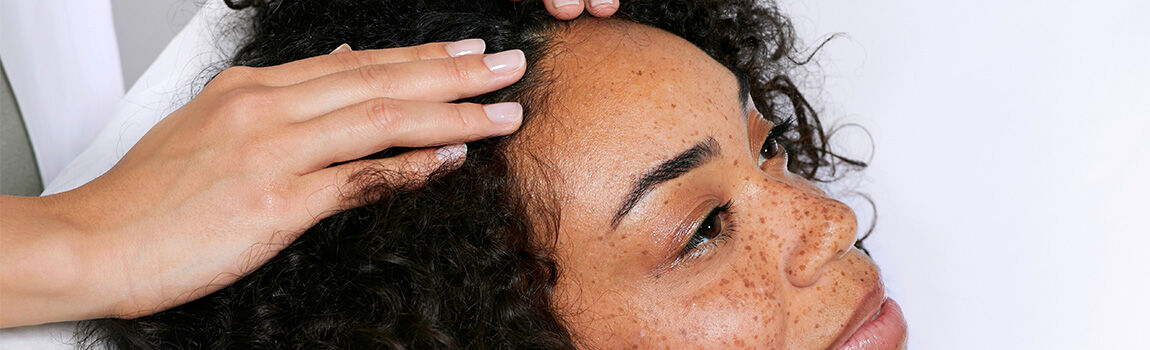 A close-up of a person with curly hair receiving a scalp massage, with a hand gently touching their forehead. The person has freckles and appears relaxed.