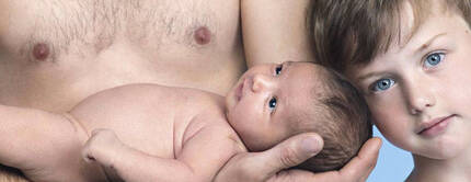 A close-up of a shirtless man holding a newborn baby in one arm, while a young boy stands closely beside them, gazing at the baby with blue eyes. The background is a soft blue.