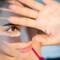 A close-up of a young woman with dark hair, wearing a pink top, shielding her eyes with her hand while looking intently at the camera, with soft shadows cast across her face.