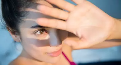A close-up of a young woman with dark hair, wearing a pink top, shielding her eyes with her hand while looking intently at the camera, with soft shadows cast across her face.