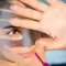 A close-up of a young woman with dark hair, wearing a pink top, shielding her eyes with her hand while looking intently at the camera, with soft shadows cast across her face.