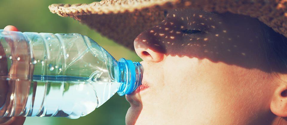 A person wearing a straw hat drinks from a plastic water bottle, with sunlight casting patterns on their face.