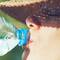 A person wearing a straw hat drinks from a plastic water bottle, with sunlight casting patterns on their face.