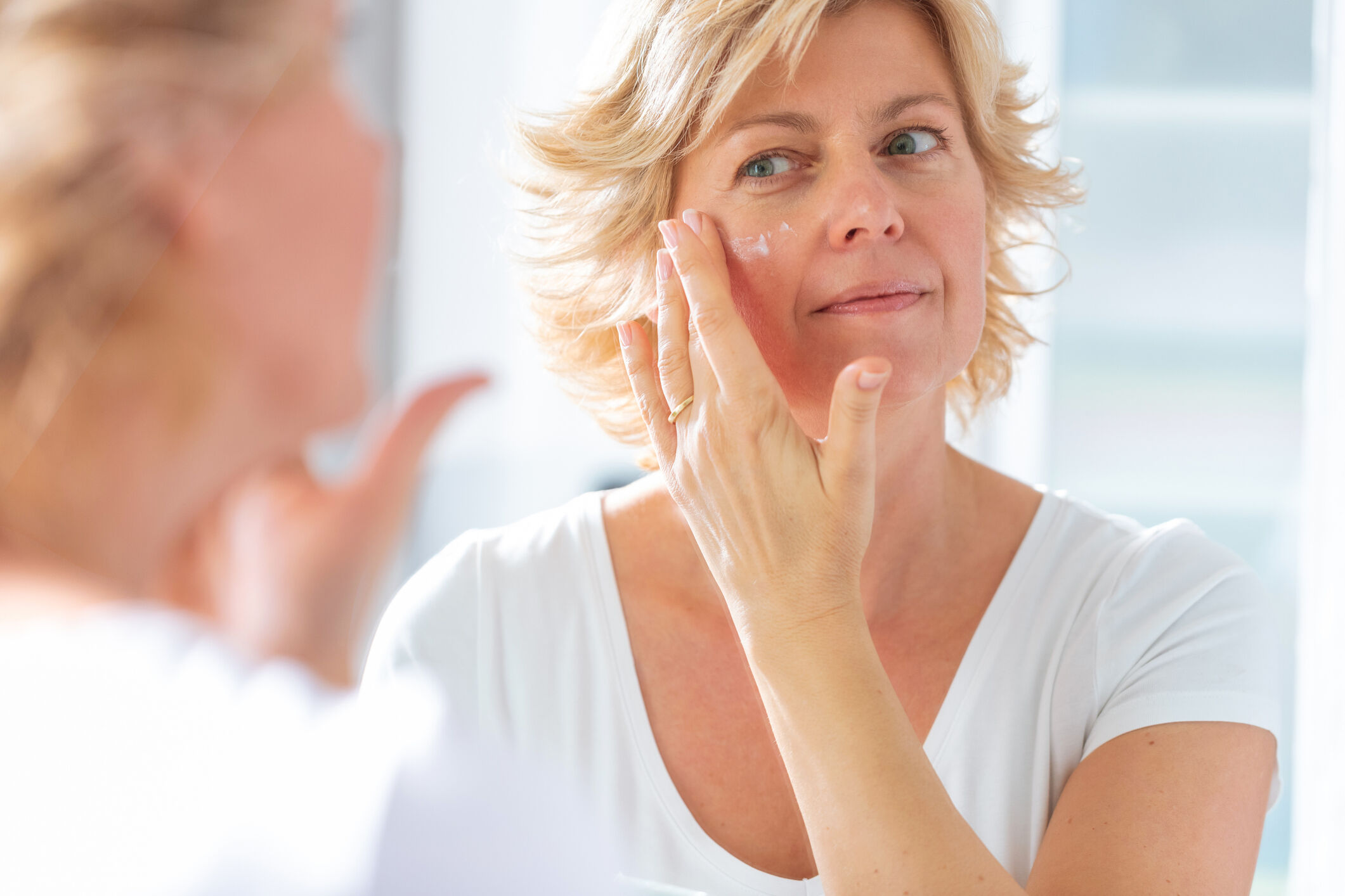 Femme mure appliquant une creme de soin sur sa joue face a son miroir pour hydrater et apaiser sa peau.