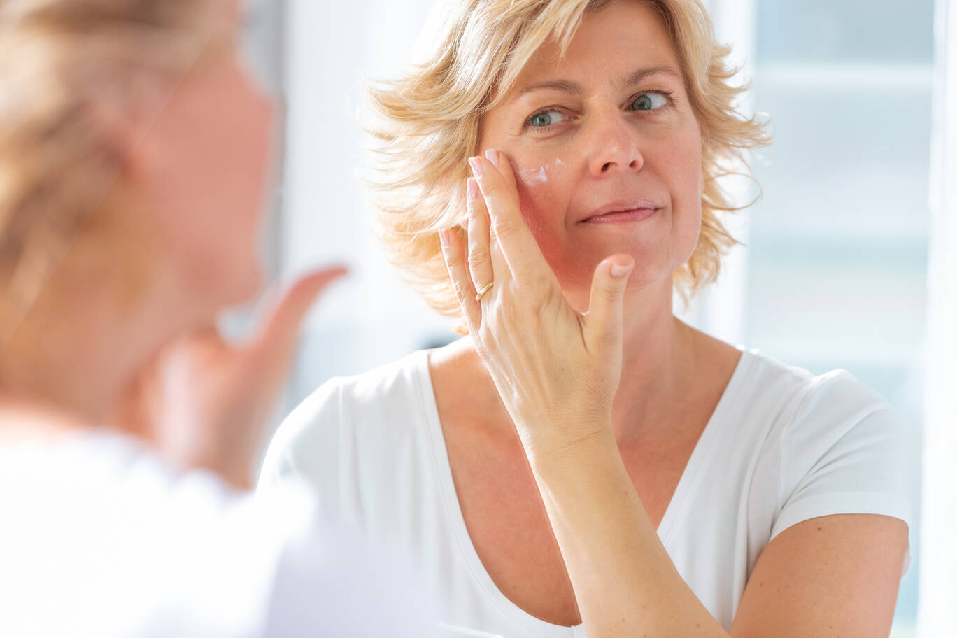Femme mure appliquant une creme de soin sur sa joue face a son miroir pour hydrater et apaiser sa peau.