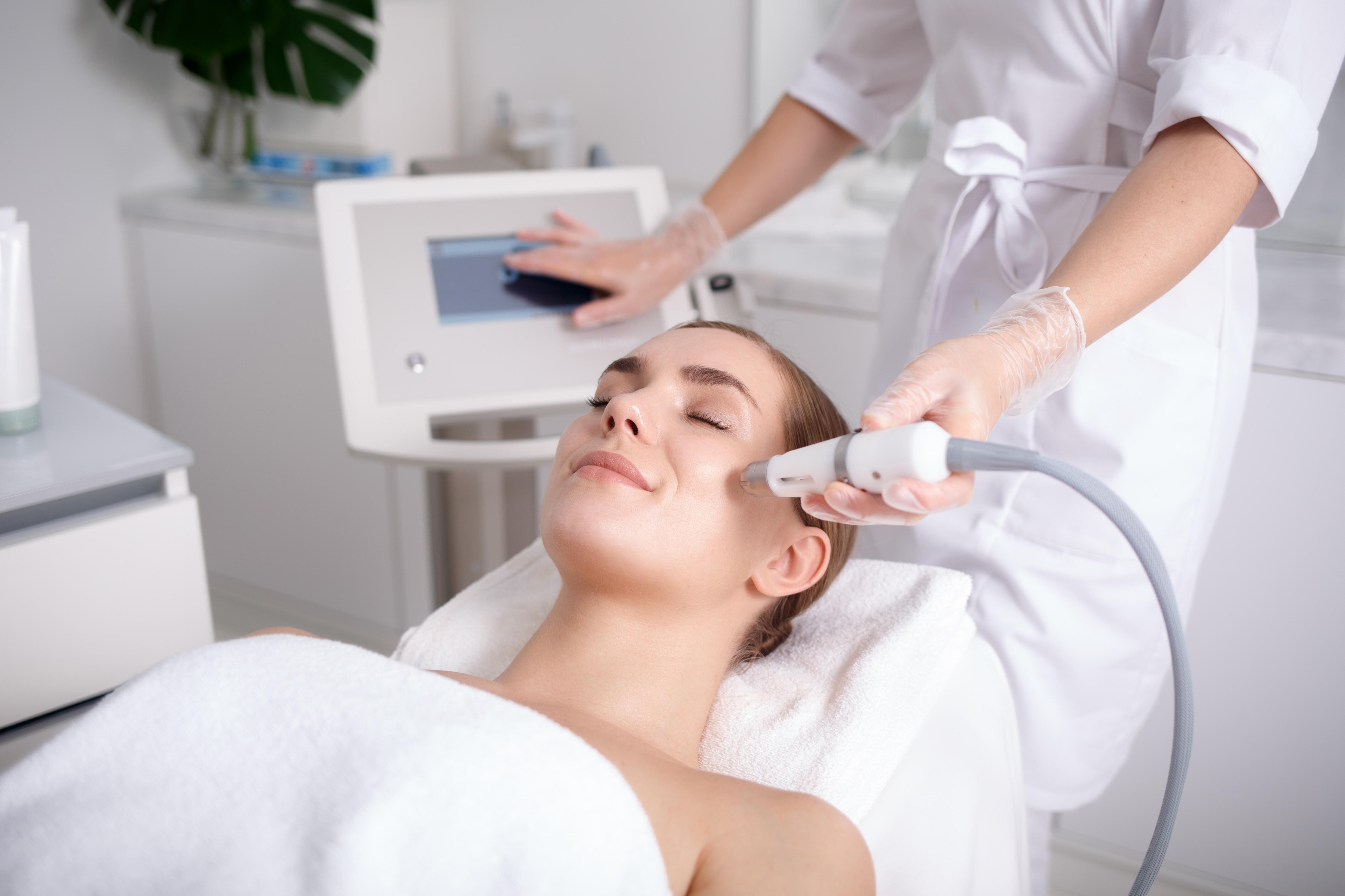 Une femme recoit un soin du visage dans un salon de beaute, tandis qu'une estheticienne utilise un appareil sur sa peau.