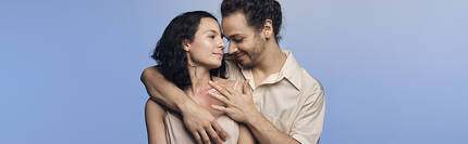Deux personnes se tiennent proches l'une de l'autre, avec un fond bleu, exprimant une connexion intime et affectueuse.
