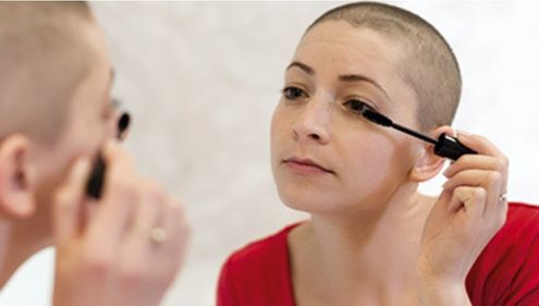 Une femme au crane rase appliquant du mascara en se regardant dans un miroir.