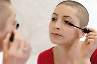 Une femme au crane rase appliquant du mascara en se regardant dans un miroir.