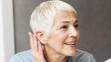 Une femme souriante aux cheveux courts argentes, touchant son oreille et regardant sur le cote.