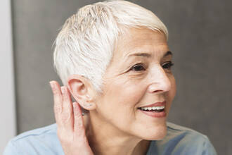 Une femme souriante aux cheveux courts argentes, touchant son oreille et regardant sur le cote.