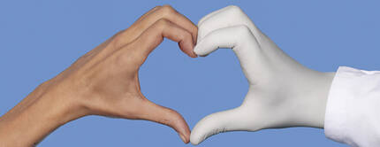 Une main nue et une main portant un gant medical blanc formant ensemble un coeur sur un fond bleu.