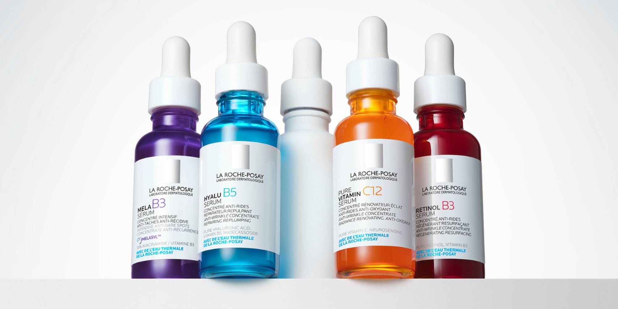 Quatre flacons de serum aux couleurs variees (violet, bleu, orange) avec pipettes, presentes sur une etagere blanche.
