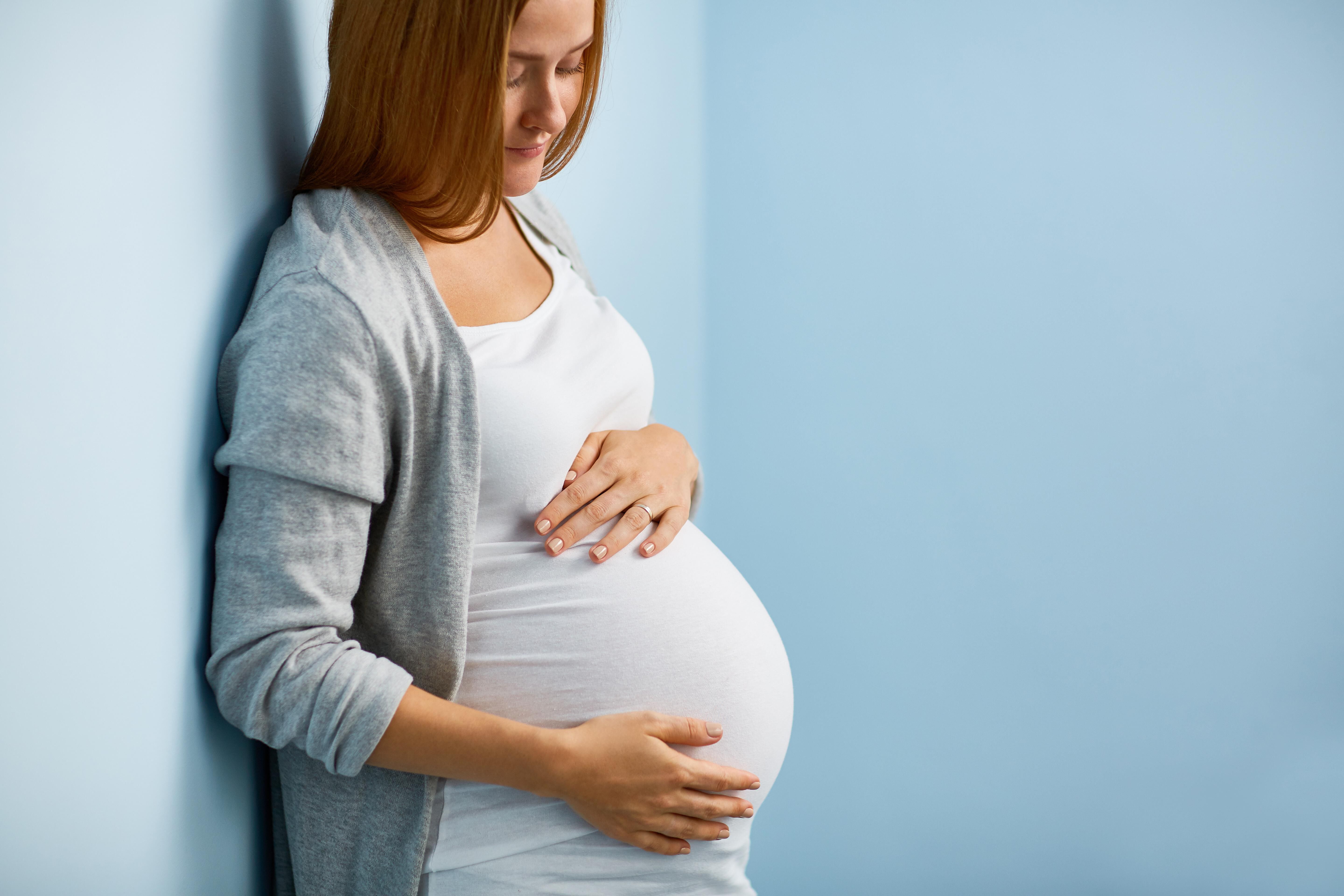 Femme enceinte, souriante, touchant son ventre arrondi, adossee a un mur bleu.