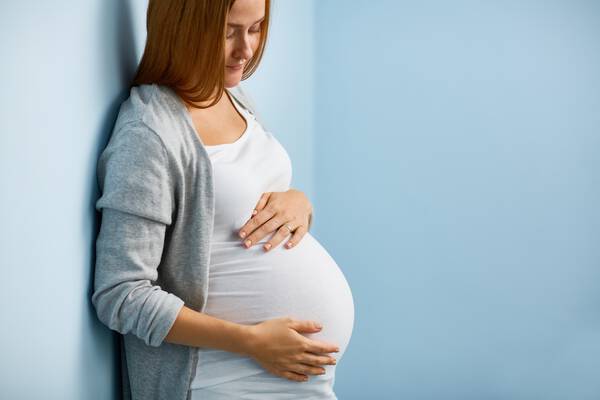 Femme enceinte, souriante, touchant son ventre arrondi, adossee a un mur bleu.