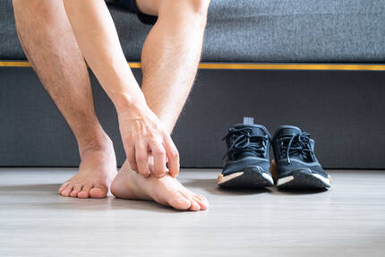 Un homme assis sur un canape se frotte la cheville, avec des chaussures de sport noires a cote de lui.