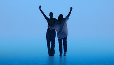 Silhouette de deux personnes s'enlacant, bras leves faisant le signe V de la victoire sur un fond bleu eclatant.