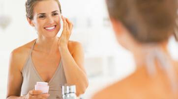 Une femme souriante s'applique une creme sur le visage devant un miroir. Elle porte un haut simple et semble satisfaite de sa routine de soins.