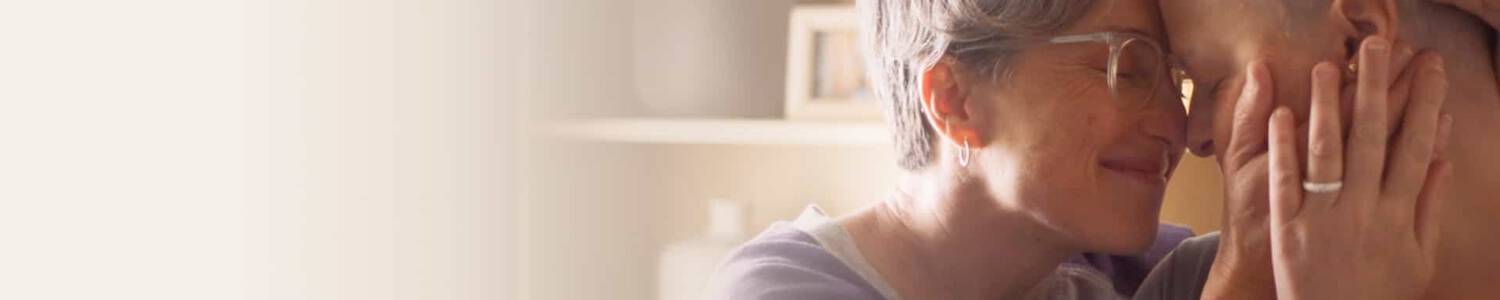 Deux personnes, d'age mur et d'age adulte, se tiennent face a face, se touchant le front avec des sourires tendres, dans un environnement chaleureux et lumineux.