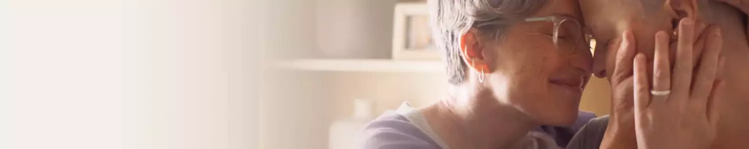 Deux personnes, d'age mur et d'age adulte, se tiennent face a face, se touchant le front avec des sourires tendres, dans un environnement chaleureux et lumineux.