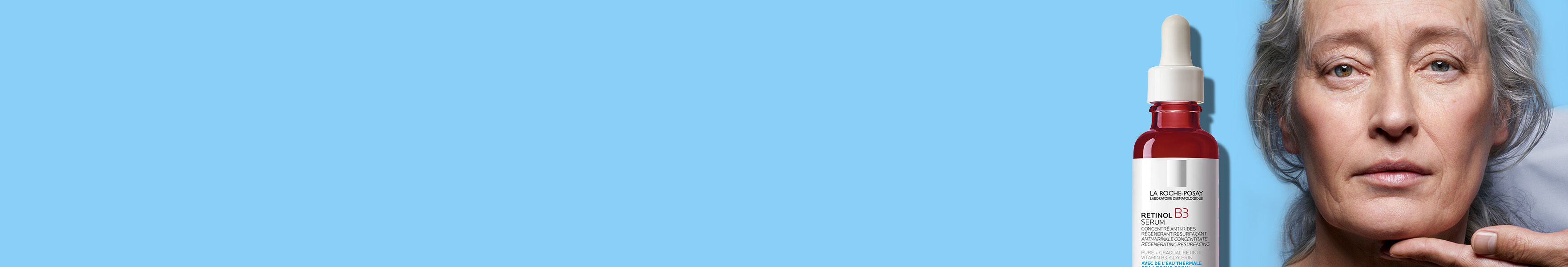 Une femme mure avec une peau lumineuse tient un flacon de serum, sur fond bleu clair.