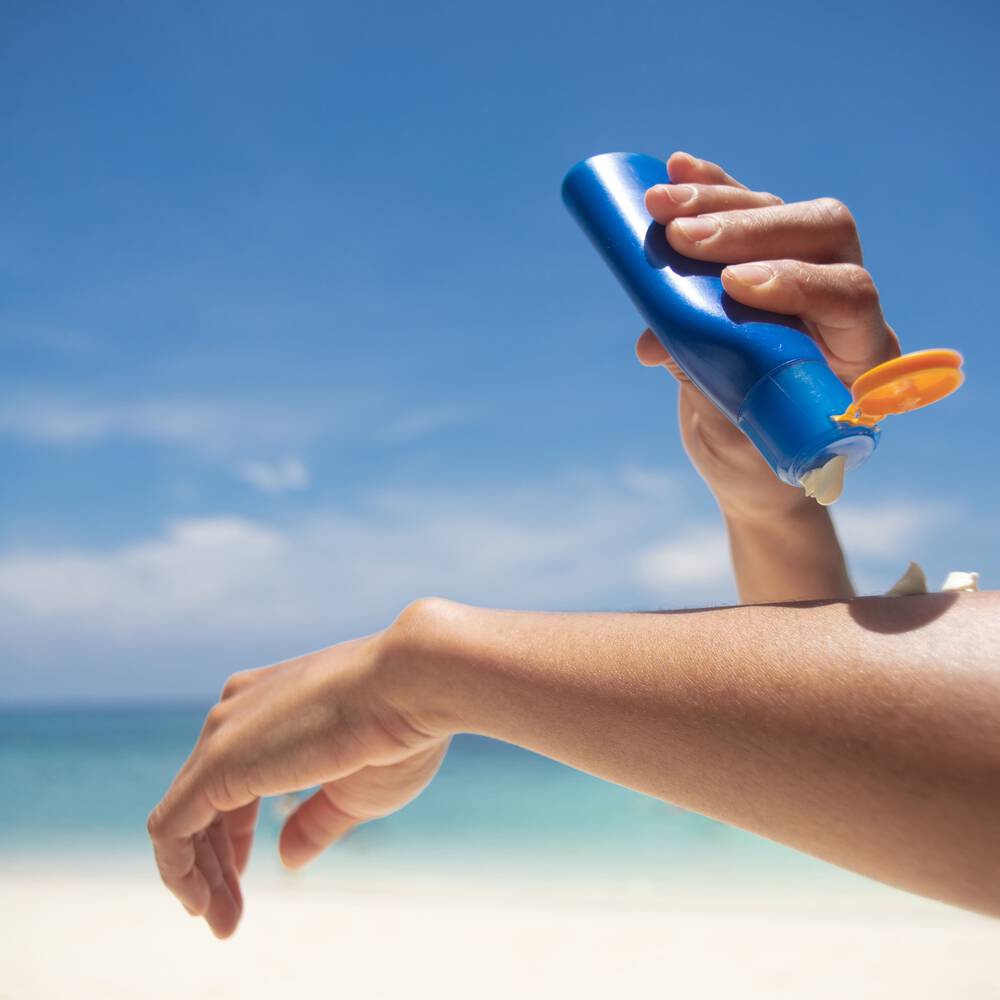 Une personne applique de la creme solaire sur son bras, avec une plage et le ciel bleu en arriere-plan.