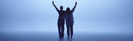 Deux personnes se tiennent debout, de dos, avec les bras leves en signe de joie, devant un fond bleu brumeux.