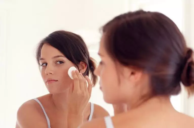 Une jeune femme s'applique du maquillage sur le visage devant un miroir, le regard concentre.