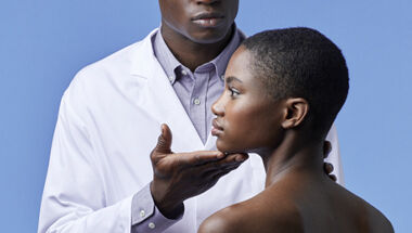Un dermatologue en blouse blanche examinant le cou et la machoire d'une jeune femme sur un fond bleu.