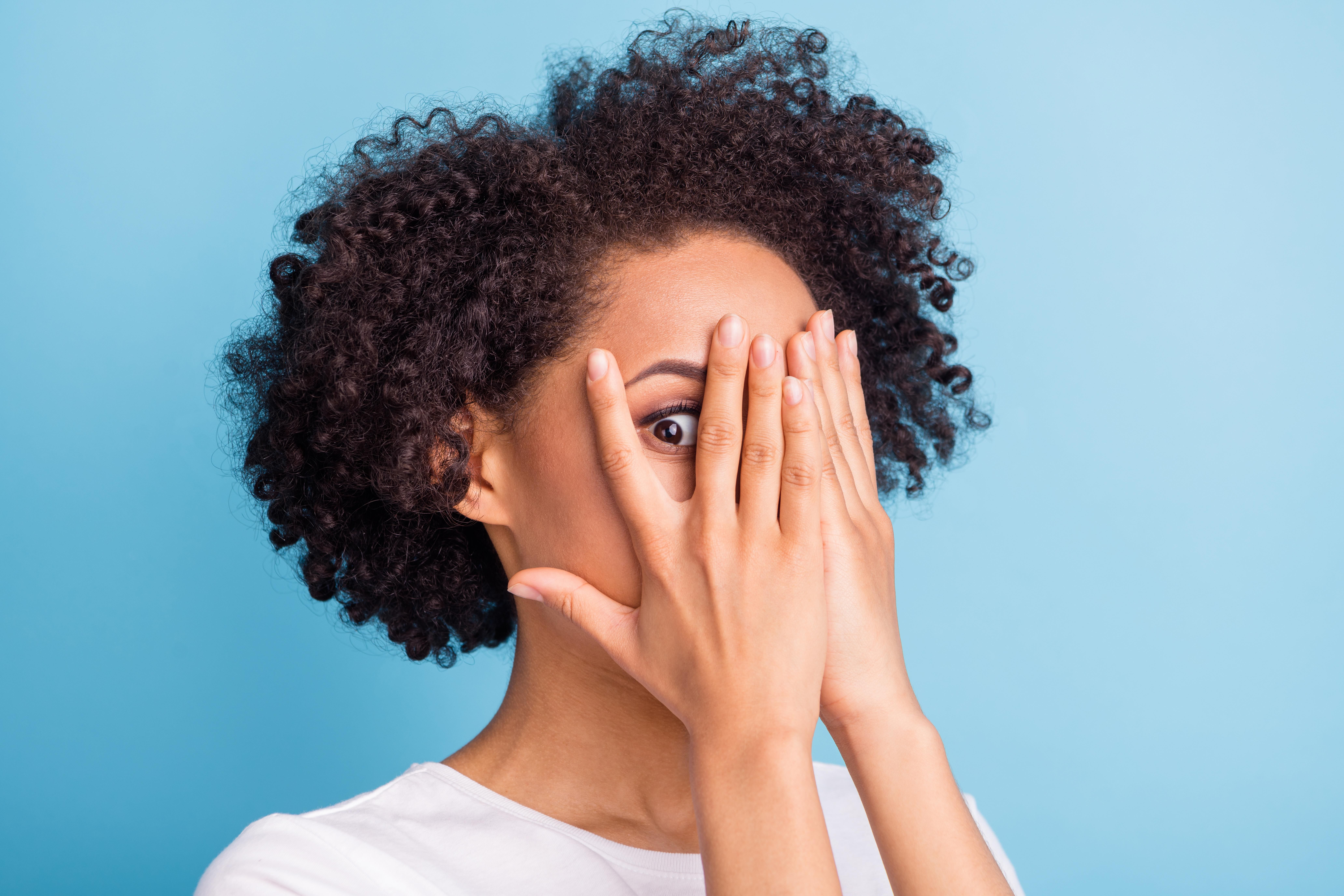 Une femme avec des cheveux crepus cachant partiellement son visage derriere ses mains, sur un fond bleu.